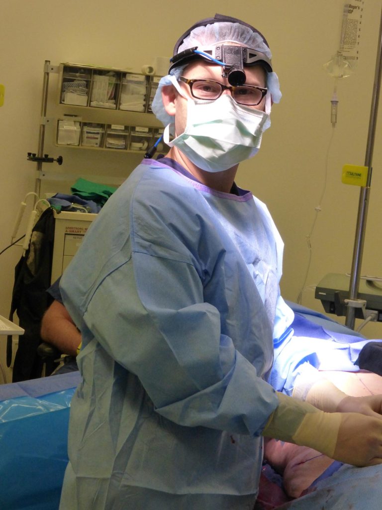 Dr. Steele performs plastic surgery while wearing his blue medical scrubs.